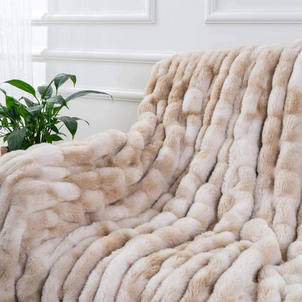 The RCL Fur Throw, in soft beige, is draped over a chair in a bright room. Nearby sits a green potted plant, and the background reveals white paneled walls.