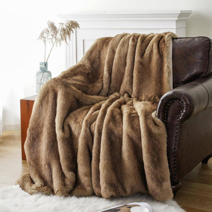 The RCL Fur Throw, a plush brown faux fur blanket, elegantly drapes over a dark leather armchair in a cozy room. A small wooden table beside the chair holds a vase with dried pampas grass, and a white rug partially covers the wooden floor.