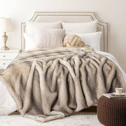 A cozy bedroom showcases the RCL Fur Throw, a reversible faux fur blanket in beige and cream, on the bed. Elegant white pillows, a decorative knotted cushion, and a nailhead trim headboard enhance the look. A nightstand with a lamp and a brown knitted ottoman complete this inviting space.