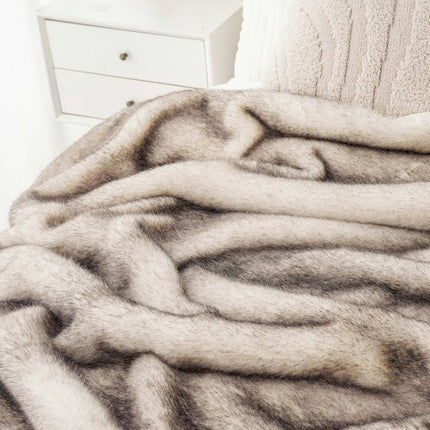 The RCL Fur Throw, a soft textured faux fur blanket in gray and beige, is elegantly draped over a bed, complementing the white nightstand with two drawers.