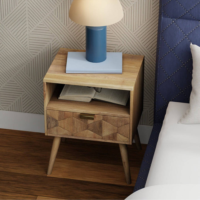 The Mango Geometric Nightstand, with its open shelf and drawer, sits beside a bed featuring a blue upholstered headboard. A lamp with a blue base and beige shade adorns it. The wooden floor enhances the bohemian flair of the geometric wall pattern.