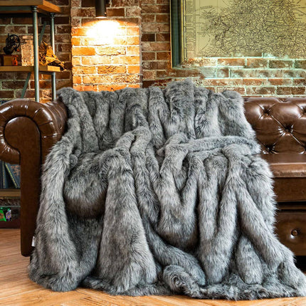 An RCL Fur Throw in plush gray faux fur elegantly drapes over a brown leather couch in a cozy room with a brick wall, adorned by a map and shelf of various items, all warmly lit by a nearby lamp.