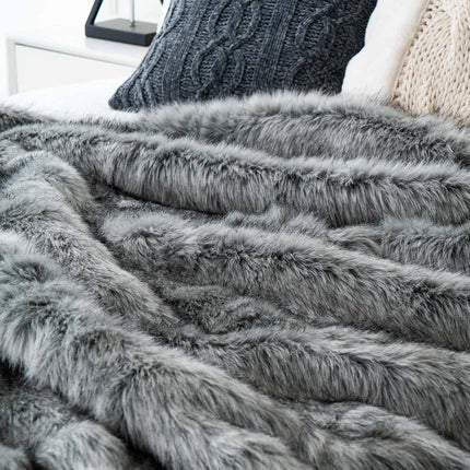 The RCL Fur Throw, a luxurious gray faux fur blanket, is draped elegantly on the bed and serves as a reversible throw. It’s paired with a dark gray cable knit pillow and a background pillow in cream knit.