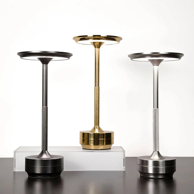 The Lumin Lamp features a modern design with circular heads and cylindrical bases in black, gold, and silver. Displayed against a white background on a black surface, the gold lamp includes a clear rectangular platform for versatile lighting options.