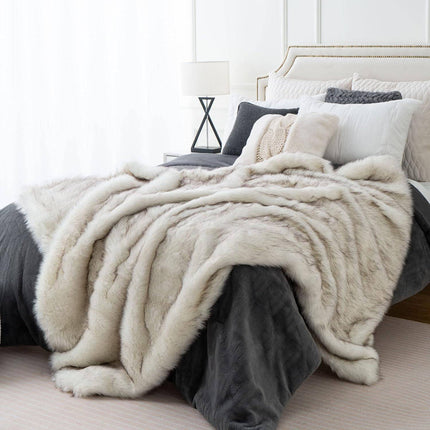 A cozy bedroom showcases a bed adorned with an RCL Fur Throw, complemented by multiple gray and white pillows. A modern lamp rests on the bedside table, and natural light streams through a large window.